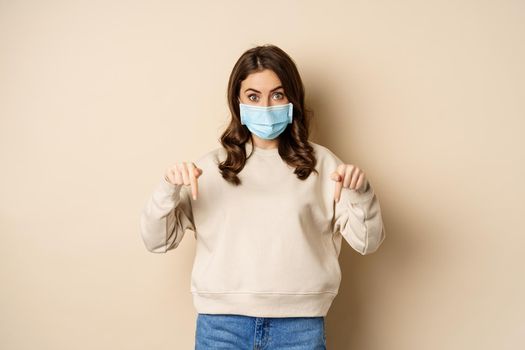 Cute girl in medical face mask, pointing fingers and looking down, showing advertisement below, standing against beige background.