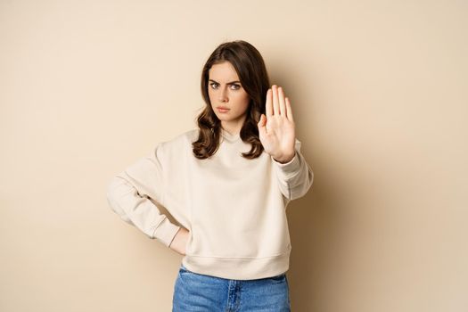 Stop. Serious and confident woman showing extended arm palm, prohibit, forbid smth, blocking something, standing over beige background.