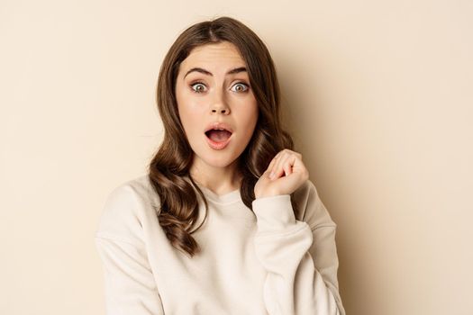 Close up shot of excited woman looking amazed with opened mouth, standing over beige background. Copy space