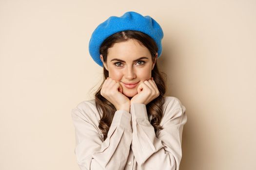 Close up portrait of feminine young woman in trendy french hat, posing cute and romantic, making silly coquettish face and tender smile, beige background.