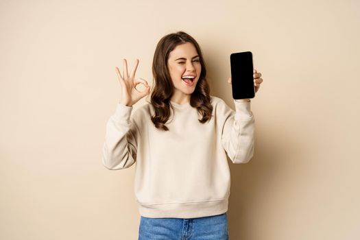 Beautiful modern woman winks, shows okay sign and mobile phone screen, app interfce, standing over beige background.