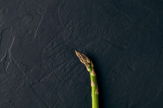 One raw fresh green asparagus stalk over black slate background. Concept of food and seasonal vegetables crop. Close up, copy space. Flat lay, top view