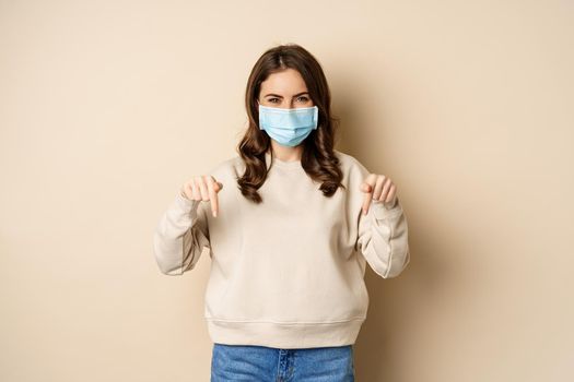 Cheerful cute woman in medical face mask, pointing fingers down, showing advertisement, using protection from covid-19, beige background.