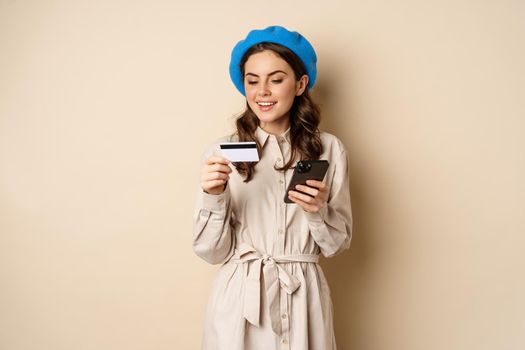 Portrait of beautiful young woman, 25 years old, using mobile phone shopping app, credit card, order delivery, purchase, standing over beige background.