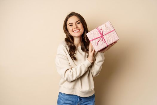 Happy beautiful girlfriend holding pink gift box and laughing, smiling joyful, concept of holidays and celebration, beige background.