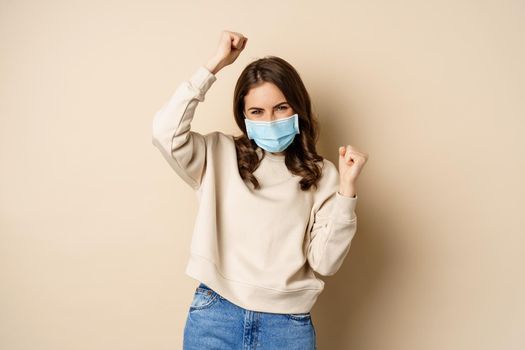 Covid-19, pandemic and quarantine concept. Enthusiastic girl dancing in face medical mask, celebrating, standing over beige background.