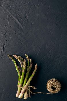 Uncooked green asparagus stems in bunch are tied with jute thread on black slate background. Skein is nearby. Food and seasonal vegetables harvest. Close up, copy space. Flat lay, top view