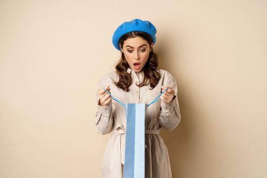 Image of girl looking surprised after opening gift bag, looking at present amazed, standing in stylish outfit over beige background.