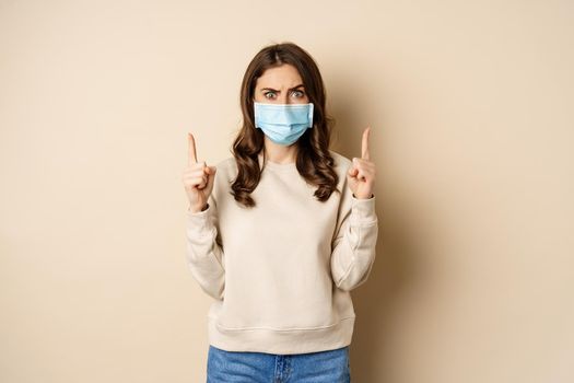 Confused woman in medical mask looking puzzled, pointing fingers up, showing smth strange on top, standing against beige background.