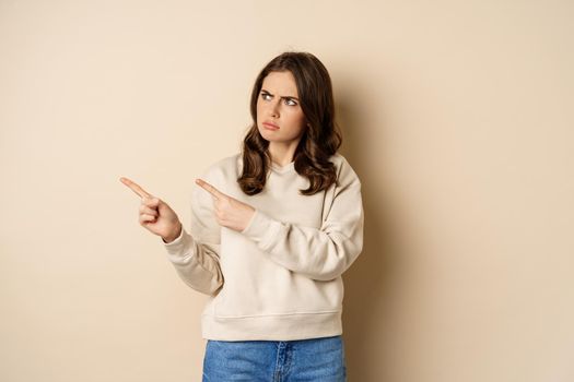 Woman pointing left and staring confused at banner copy space, strange logo, standing over beige background.