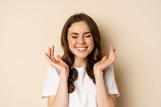 Close up portrait of enthusiastic young woman rejoicing, shouting with joy and satisfaction, celebrating victory, winning and triumphing, standing over beige background.