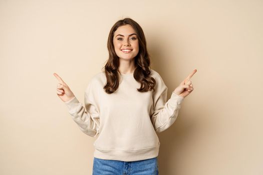 Cheerful caucasian woman showing directions, two ways, pointing sideways at variants, choices for customer, standing over beige background.