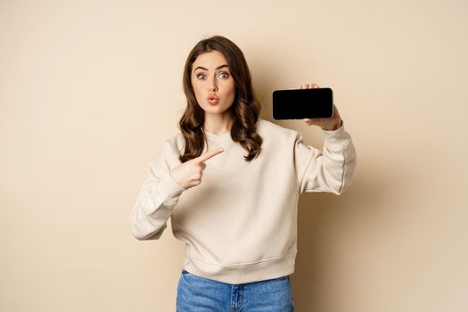 Image of girl pointing finger, showing mobile phone horizontal screen and looking amazed by application, standing over beige background.