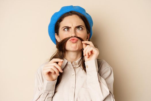 Funny beautiful girl fooling around, making moustache out of hair under lips, squinting and grimacing, standing happy against beige background.