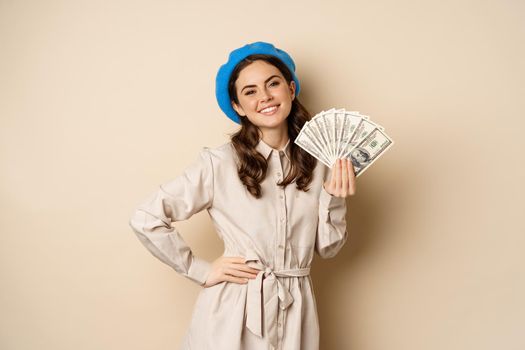 Stylish young woman holding money, cash dollars, smiling and posing satisfied, going on shopping, standing over beige background.