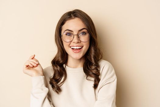 Close up portrait of stylish brunette woman in glasses, laughing and smiling, posing in eyewear against beige background.