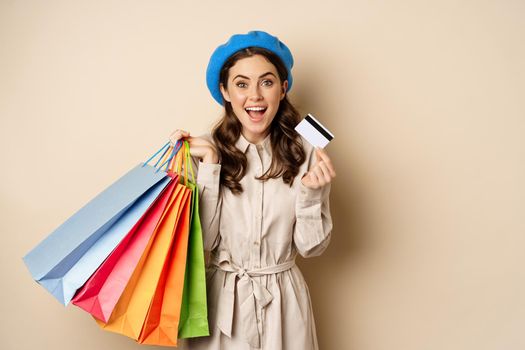 Portrat of trendy feminine girl posing with shopping bags from store and credit card, paying contactless, buying with discount on sale, beige background.