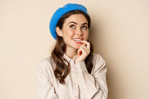 Close up portrait of dreamy smiling girl imaging, picturing smth, looking up and thinking, standing over beige background in blue trendy hat.