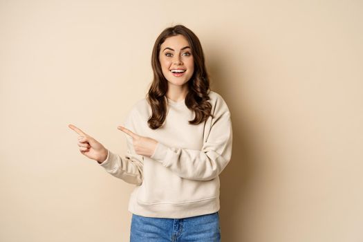 Cheerful attractive female model pointing fingers left, showing store advertisement, banner or logo, standing in sweater over beige background.