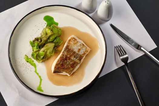 Delicious fish cooked in an oven and garnished with sauce and cabbage, served on a white plate with black edging, dark table, cutlery. Selective focus. Top view, restaurant cooking concept.