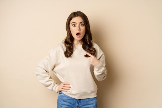 Surprised girl pointing at herself and look with disbelief, standing over beige background.