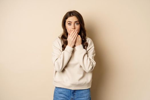 Shocked and scared woman cover mouth, looking stunned and speechless, worry about smth, standing over beige background.