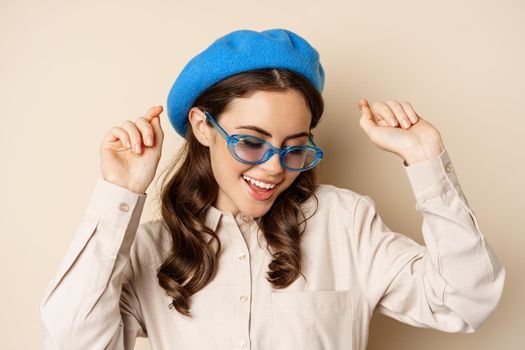 Portrait of stylish happy woman dancing in trendy sunglasses, standing over beige studio background.