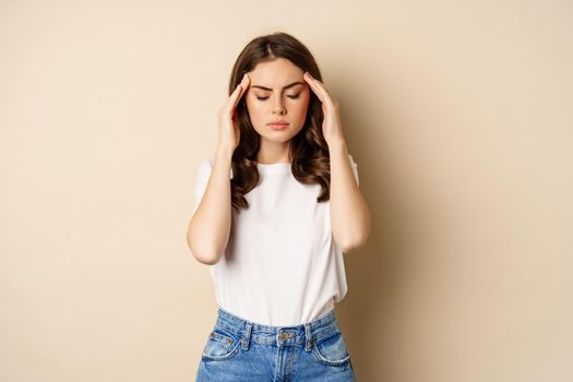 Health and women concept. Portrait of girl feeling sick, touching head temples, feeling headache, migraine, standing over beige background.