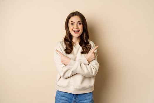 Happy smiling girl brunette pointing sideways, showing two promo offers, choice in store, standing over beige background.