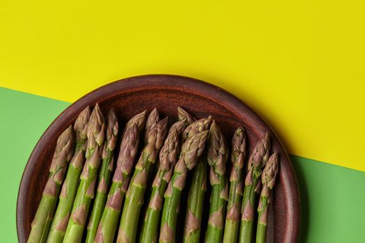 Raw ripe asparagus stalks in a brown clay plate, over yellow and green background. Concept of food and seasonal vegetables harvest. Close up, copy space. Flat lay, top view