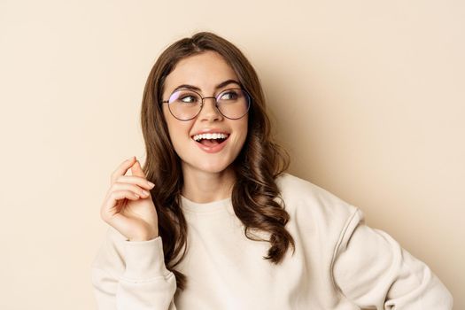 Stylish young caucasian woman wearing glasses and smiling, posing against beige background. Copy space