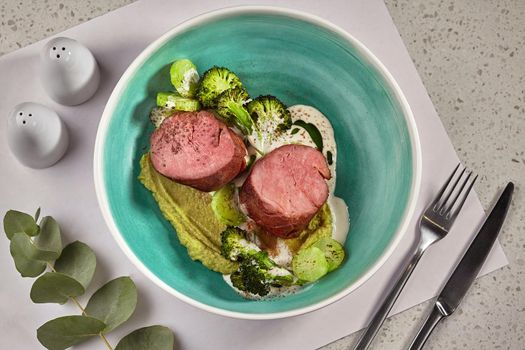 Close-up photo of a delicious roasted meat with broccoli and zucchini seasoned with sauce, decorated with a green twig. Served on a blue deep plate, cutlery. Restaurant cooking concept. Top view.