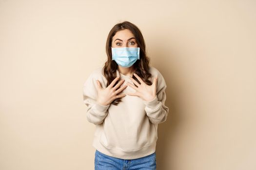 Covid-19, pandemic and quarantine concept. Surprised brunette woman in face medical mask, looking amazed at camera, watching big news, beige background.