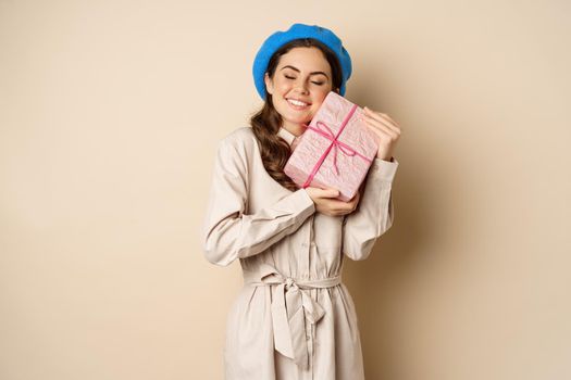 Holidays and gifts concept. Beautiful girl receive gift box and looking happy, holding pink wrapped present with joyful face expression, beige background.