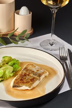 Tasty fish cooked in an oven and garnished with sauce and cabbage, served on a white plate with black edging, glass of wine, dark table, cutlery. Selective focus. Top view, restaurant cooking concept.