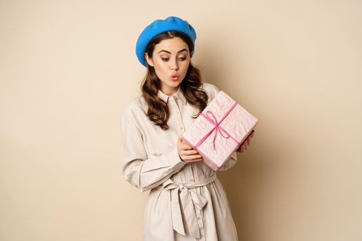 Holidays and gifts concept. Beautiful girl receive gift box and looking happy, holding pink wrapped present with joyful face expression, beige background.