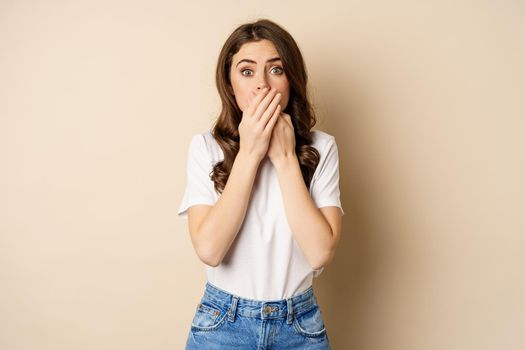 Shocked brunette woman gasping, looking speechless at camera, cover mouth with hands, standing over beige background.