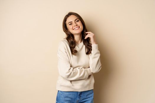 Beautiful young brunette woman posing happy against beige background, smiling at camera, wearing sweater.