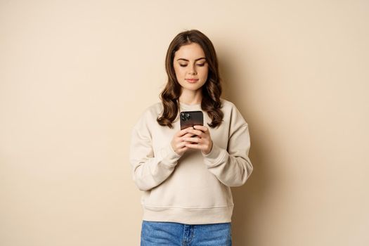 People and cellular technology. Beautiful stylish woman using mobile phone, smartphone app, smiling and looking at screen, standing over beige background.