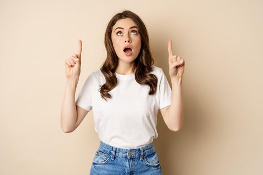 Surprised brunette girl showing promo, pointing and looking up with amazed face expression, standing over beige background.