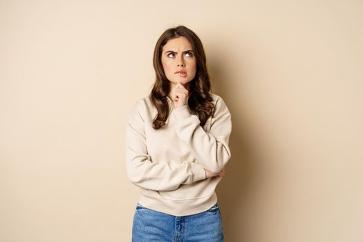 Image of beautiful woman with serious face thinking, standing over beige background.