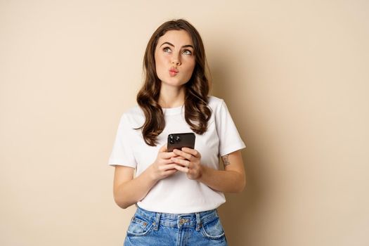 Portrait of woman thinking, holding mobile phone and looking up with thoughtful face, standing over beige background. Copy space