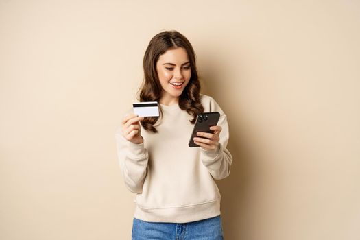 Smiling girl using mobile app, smartphone shopping and credit card, standing over beige background, order smth.