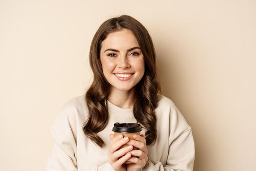 Beautiful authentic woman smiling, holding warm cup of coffee and looking happy, standing over beige background.