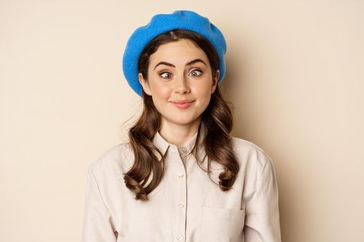 Close up portrait, silly face of young funny girl, squinting eyes and having fun, posing in trench and hat against beige background.