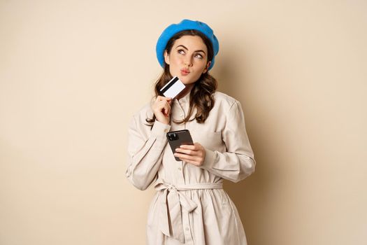 Portrait of trendy woman in coat holding smartphone, showing credit card, shopping online, order something on mobile phone, standing over beige background.