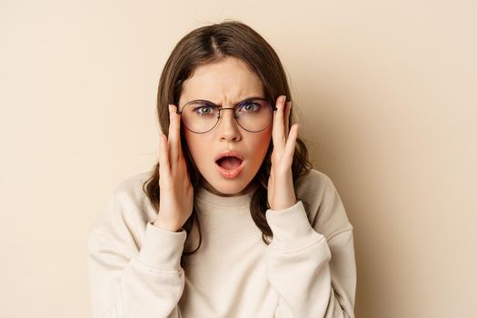 Close up portrait of woman in glasses looking confused and frustrated, cant understand smth strange, standing over beige background.