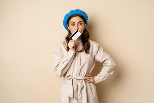 Shopping and discounts concept. Young stylish modern girl, posing with credit card, looking happy and smiling, standing over beige background.