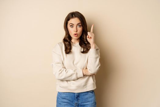Young woman suggests an idea, raising finger, has revelation, pitching a plan, pointing up, standing over beige background.