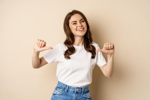 Self-assured young happy woman pointing fingers at herself and dancing, self-promoting, being confident, standing over beige background.